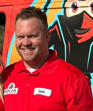 Photograph of the owner of Ninja Plumbing, Heating and Air. He is wearing a red polo shirt with the ninja logo, and his name patched onto it. The background is a Ninja HVAC company truck.